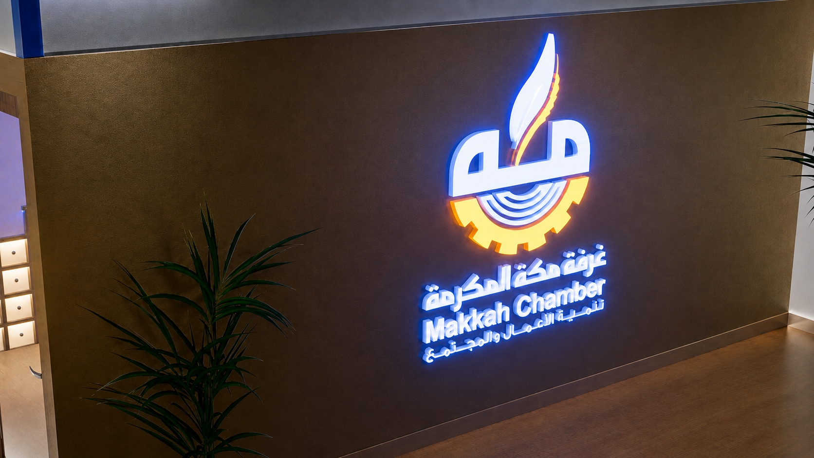 Illuminated Makkah Chamber logo and bilingual signage mounted on a warm metallic exhibition wall beside integrated display storage.