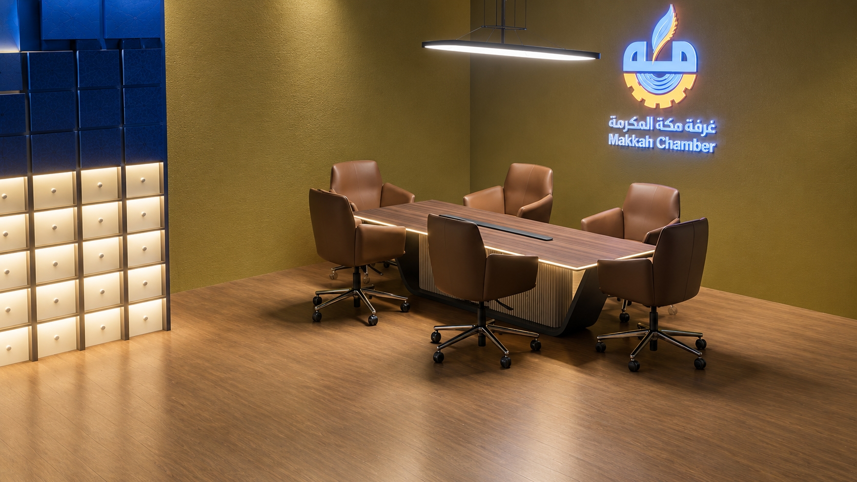 Makkah Chamber meeting room and storage area with an illuminated conference table, warm metallic walls, blue display storage, and branded signage.